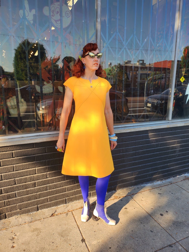 May include: A woman wearing a yellow dress, blue tights, and white shoes. She is standing in front of a shop with a window display.
