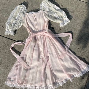 May include: A pale pink dress with a lace bodice and sleeves. The dress has a gathered skirt and a lace trim around the hem. The dress is tied at the waist with a pink ribbon.