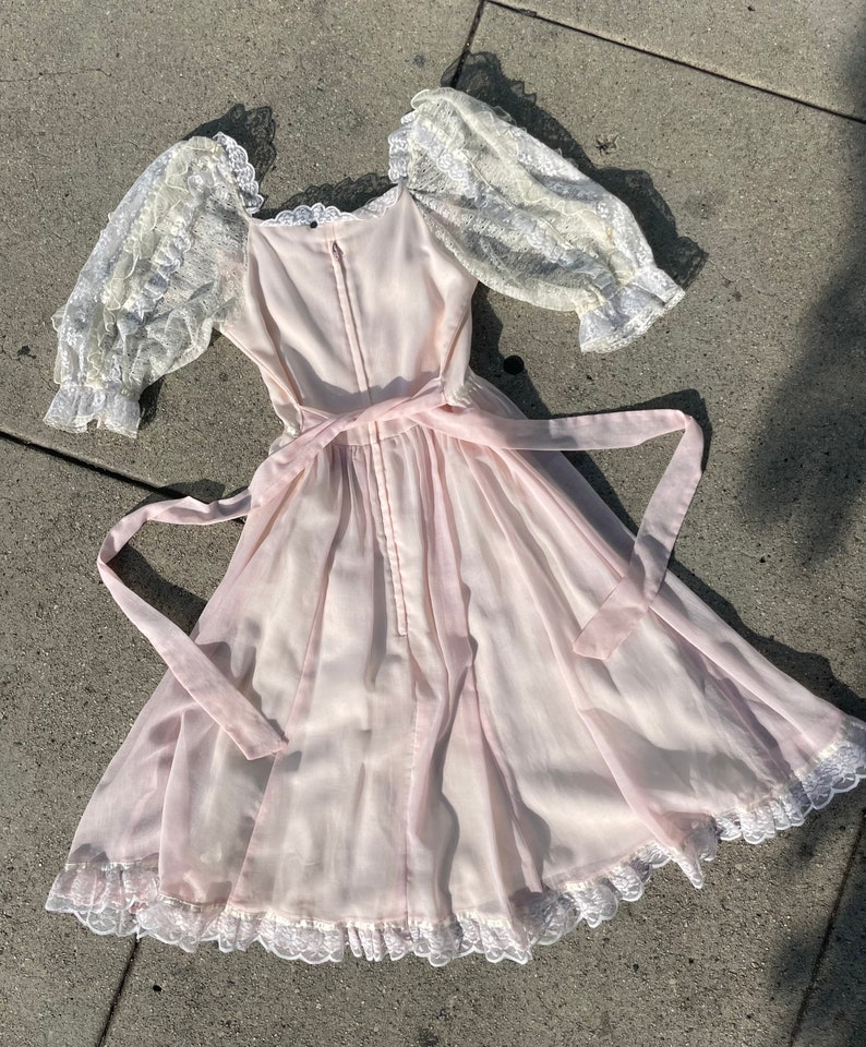 May include: A pale pink dress with a lace bodice and sleeves. The dress has a gathered skirt and a lace trim around the hem. The dress is tied at the waist with a pink ribbon.