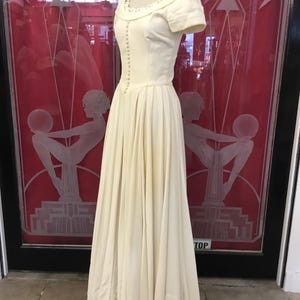 May include: An ivory-colored vintage dress displayed on a mannequin. The dress features a fitted bodice with small buttons, a scoop neckline with delicate trim, and short sleeves. The full skirt falls to the floor. The background includes red doors with art deco style etched glass.