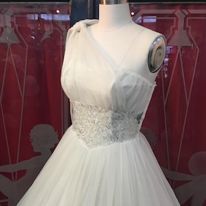 May include: A white wedding dress on a mannequin. The dress features a one-shoulder design with a flowing tulle skirt. The bodice is embellished with silver beading. The background includes a red wall with a decorative design.