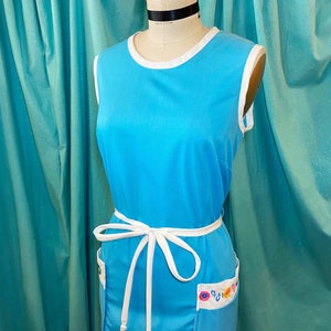 May include: A sleeveless blue dress with a white trim around the neckline and a white ribbon tied at the waist. The dress has a floral pattern on the bottom hem.