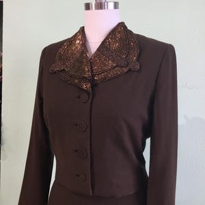 May include: A dark brown vintage suit with a beaded collar. The jacket has a button-up front and long sleeves. The collar is embellished with gold-colored beads. The suit is displayed on a mannequin.