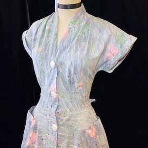 May include: A light blue dress with a floral print featuring pink flowers. The dress has a short sleeve, a button-up front, and a fitted bodice.