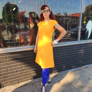 May include: A woman wearing a bright yellow dress with a short sleeve and a scoop neck. She is wearing blue tights and white boots with a clear heel. She is standing in front of a shop front with a brick wall.