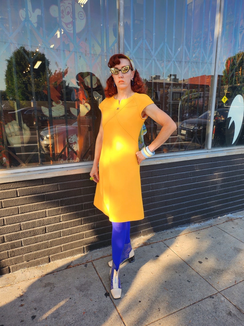 May include: A woman wearing a bright yellow dress with a short sleeve and a scoop neck. She is wearing blue tights and white boots with a clear heel. She is standing in front of a shop front with a brick wall.