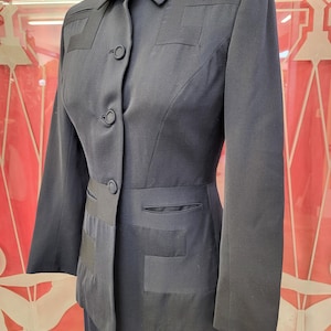 May include: A dark navy blue blazer featuring a structured collar, long sleeves, and a front button closure. The garment is detailed with tonal geometric fabric panels on the chest and waist, along with two horizontal slit pockets at the waist.