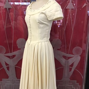 May include: A vintage ivory-colored dress displayed on a mannequin. The dress features a fitted bodice with small buttons, short sleeves, and a pleated skirt. The neckline is adorned with delicate lace trim. The dress is displayed in front of a red background.