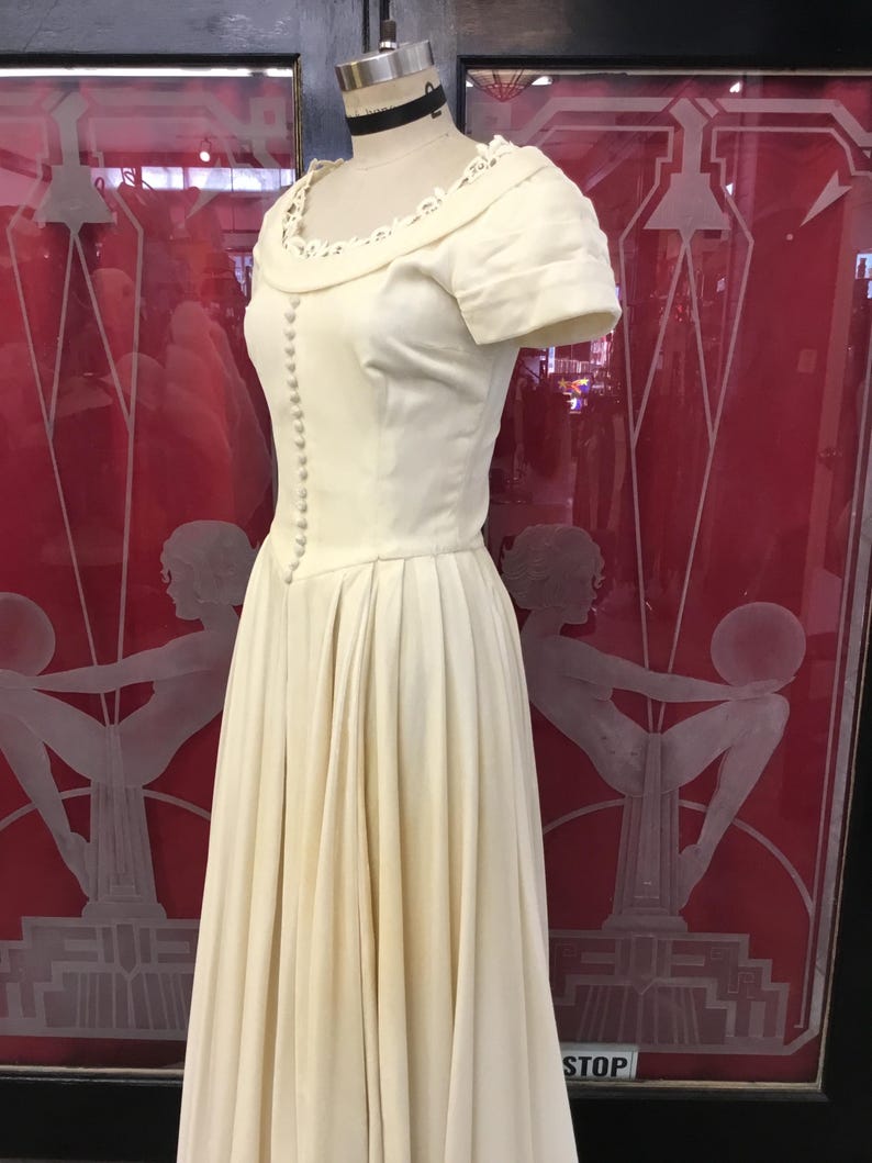May include: A vintage ivory-colored dress displayed on a mannequin. The dress features a fitted bodice with small buttons, short sleeves, and a pleated skirt. The neckline is adorned with delicate lace trim. The dress is displayed in front of a red background.
