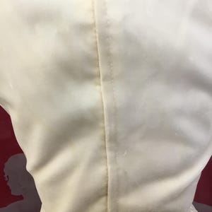 May include: Close-up of a cream-colored dress, showcasing the back with a central seam. The fabric appears to be a soft, possibly velvet or satin material. The dress has a simple, elegant design.