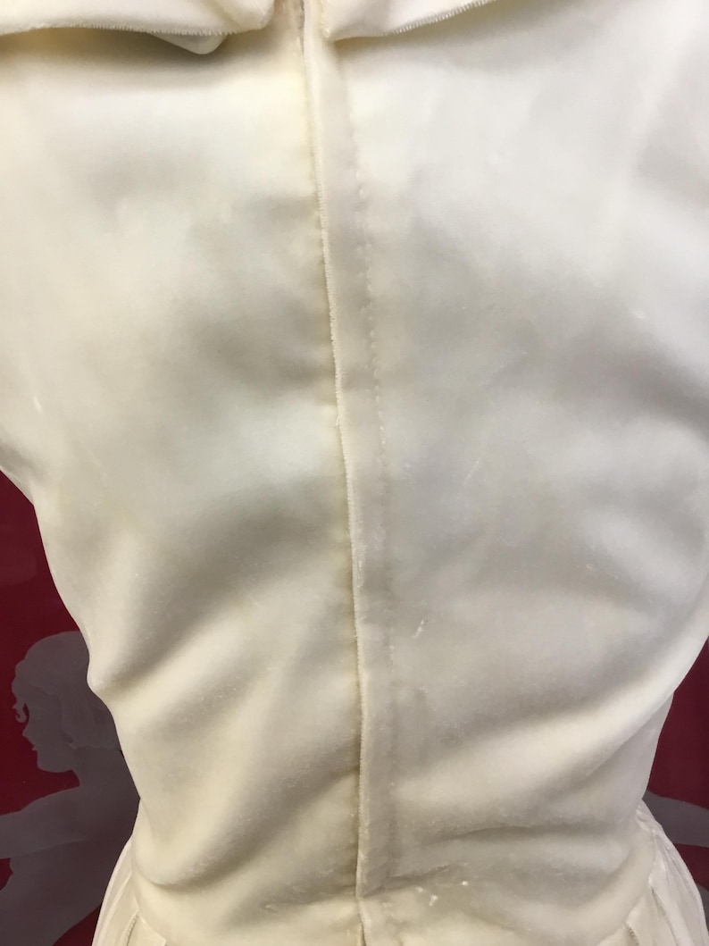 May include: Close-up of a cream-colored dress, showcasing the back with a central seam. The fabric appears to be a soft, possibly velvet or satin material. The dress has a simple, elegant design.