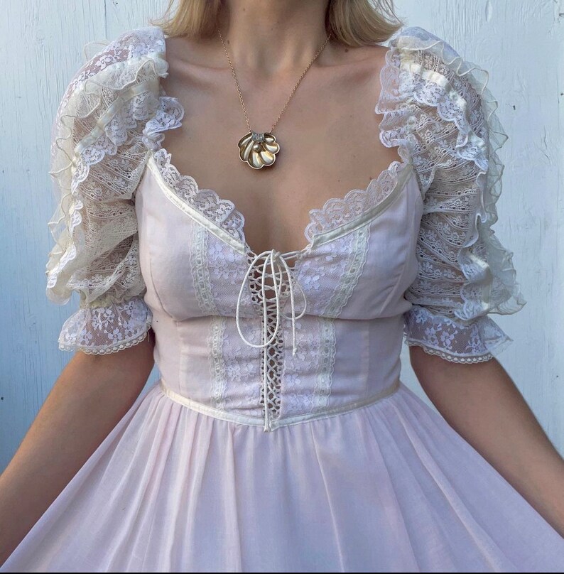 May include: A light pink dress with a lace bodice and ruffled sleeves. The dress has a lace-up detail in the front and a full skirt.