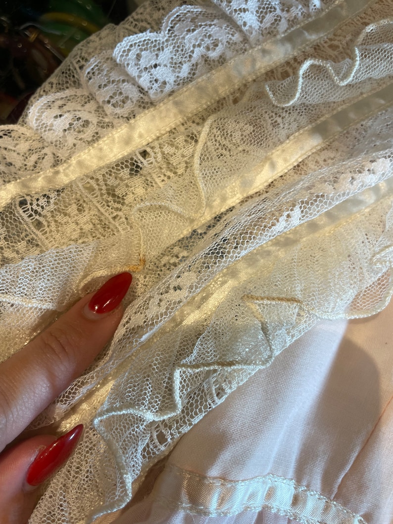 May include: Close-up of a white lace trim with a satin ribbon. The lace has a delicate floral pattern and is layered with ruffles.
