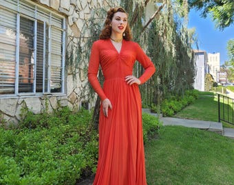 May include: A woman wearing a long, red, pleated dress with a V-neck and long sleeves. She is standing in front of a stone building with a green lawn and trees in the background.