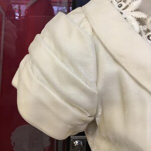 May include: Ivory-colored velvet sleeve of a garment, featuring a gathered, puffed design. The sleeve is detailed with subtle stitching and is part of a larger piece, possibly a dress or jacket. The collar is visible, with delicate lace trim.