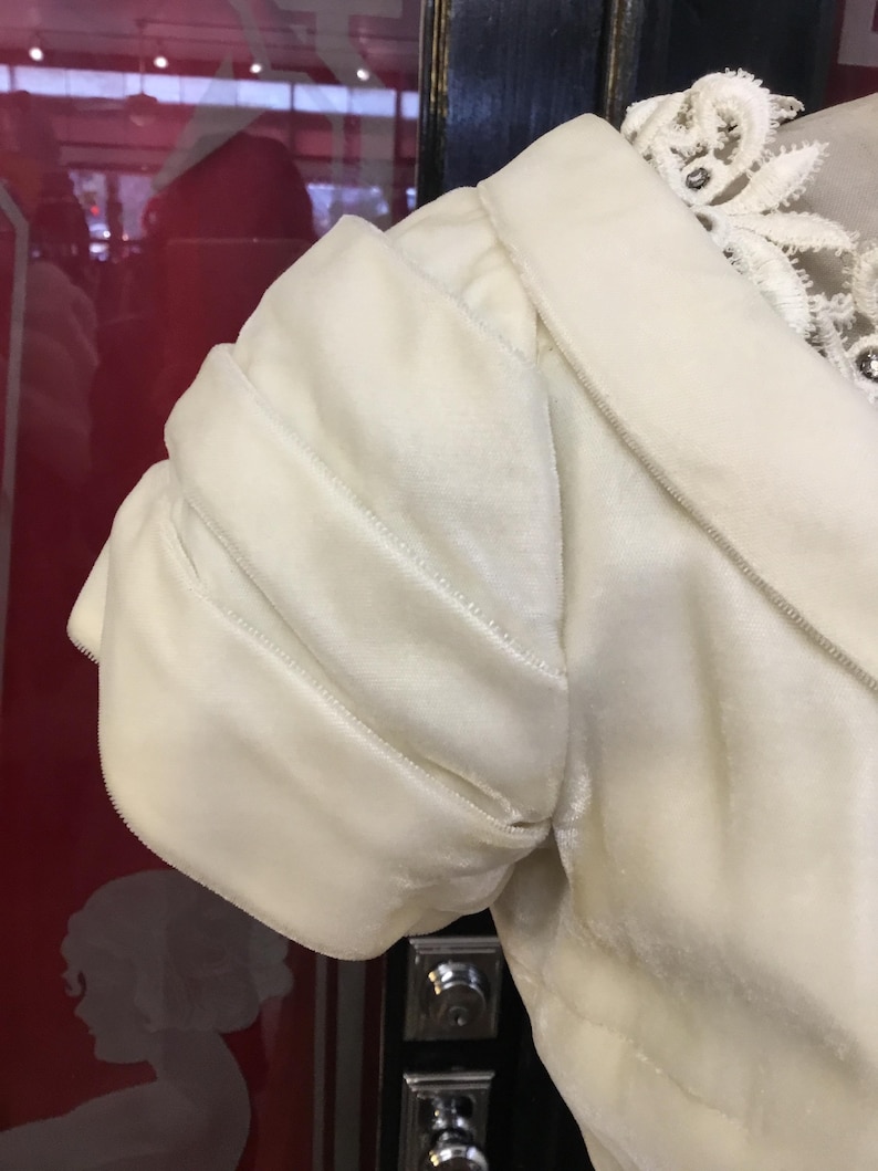 May include: Ivory-colored velvet sleeve of a garment, featuring a gathered, puffed design. The sleeve is detailed with subtle stitching and is part of a larger piece, possibly a dress or jacket. The collar is visible, with delicate lace trim.