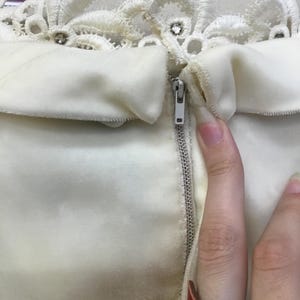 May include: Ivory-colored dress with a decorative lace collar adorned with rhinestones. The dress features a zipper closure and a soft, velvety texture. The image focuses on the upper portion of the garment.
