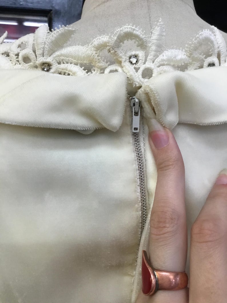May include: Ivory-colored dress with a decorative lace collar adorned with rhinestones. The dress features a zipper closure and a soft, velvety texture. The image focuses on the upper portion of the garment.