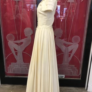 May include: Cream-colored vintage dress with a fitted bodice and a flowing pleated skirt. The dress features short sleeves, a row of buttons down the front, and a small collar. The dress is displayed on a mannequin in front of a red door.