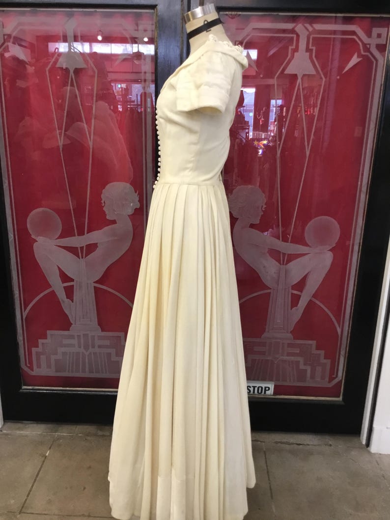 May include: Cream-colored vintage dress with a fitted bodice and a flowing pleated skirt. The dress features short sleeves, a row of buttons down the front, and a small collar. The dress is displayed on a mannequin in front of a red door.