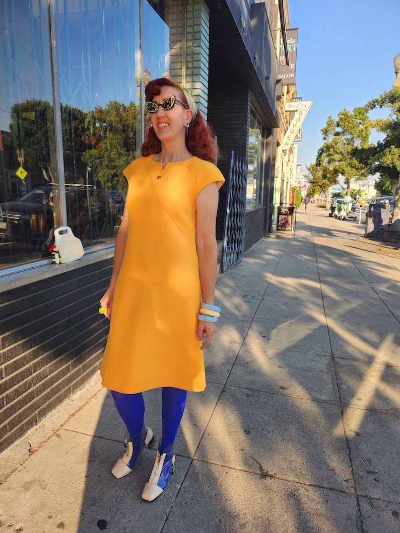 May include: A woman wearing a bright yellow dress with a blue and white patterned headband, blue tights, and white shoes. She is standing on a pavement in front of a building with a sign that says "Smash Burger".