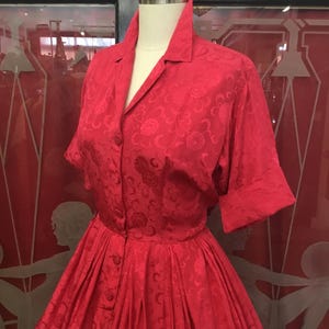 May include: A vibrant red dress with a floral pattern, displayed on a mannequin. The dress features a collared neckline, button-down front, and short sleeves. The skirt is full and pleated, creating a classic vintage silhouette.