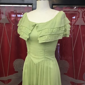 May include: A light green vintage dress on a mannequin. The dress features a fitted bodice, a flowing skirt, and a ruffled overlay with lace trim. The dress is a soft, pastel green color.