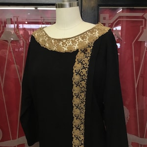 May include: Black dress with long sleeves, featuring a gold lace trim along the neckline and a vertical panel down the front. The dress is displayed on a mannequin, showcasing its design and fit. The lace detailing adds a touch of elegance to the garment.