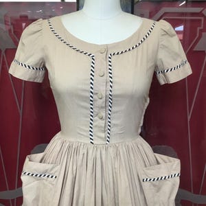 May include: A beige dress with short sleeves, a round neckline, and a button-down front. The garment features black and white striped piping along the neckline, sleeve cuffs, front placket, and the top edges of the two large side pockets.