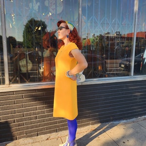 May include: A woman wearing a yellow dress, blue tights, and white shoes with blue accents. She is standing in front of a shop front with a large window.