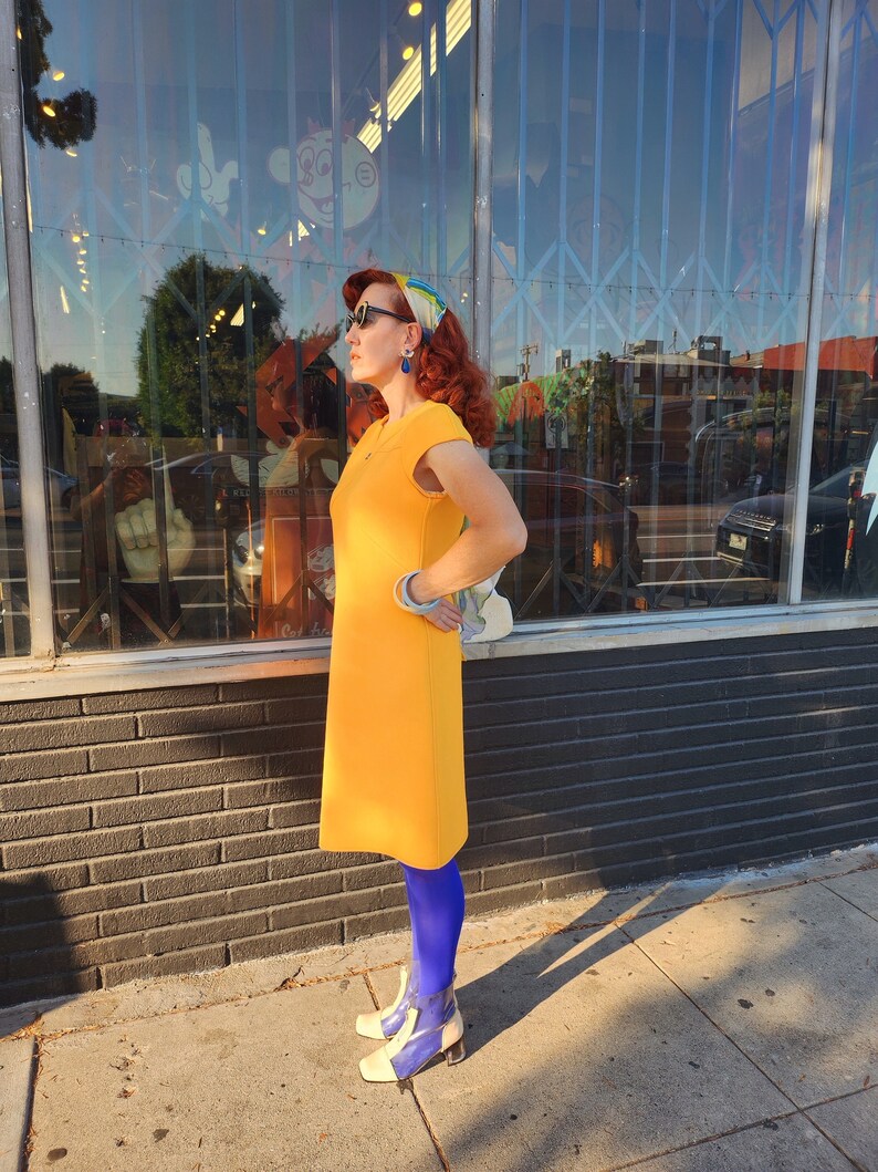 May include: A woman wearing a yellow dress, blue tights, and white shoes with blue accents. She is standing in front of a shop front with a large window.