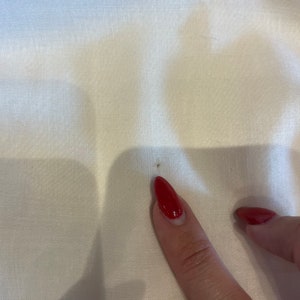 May include: A close-up of a white fabric surface with a small brown stain. A finger with red nail polish is pointing at the stain.