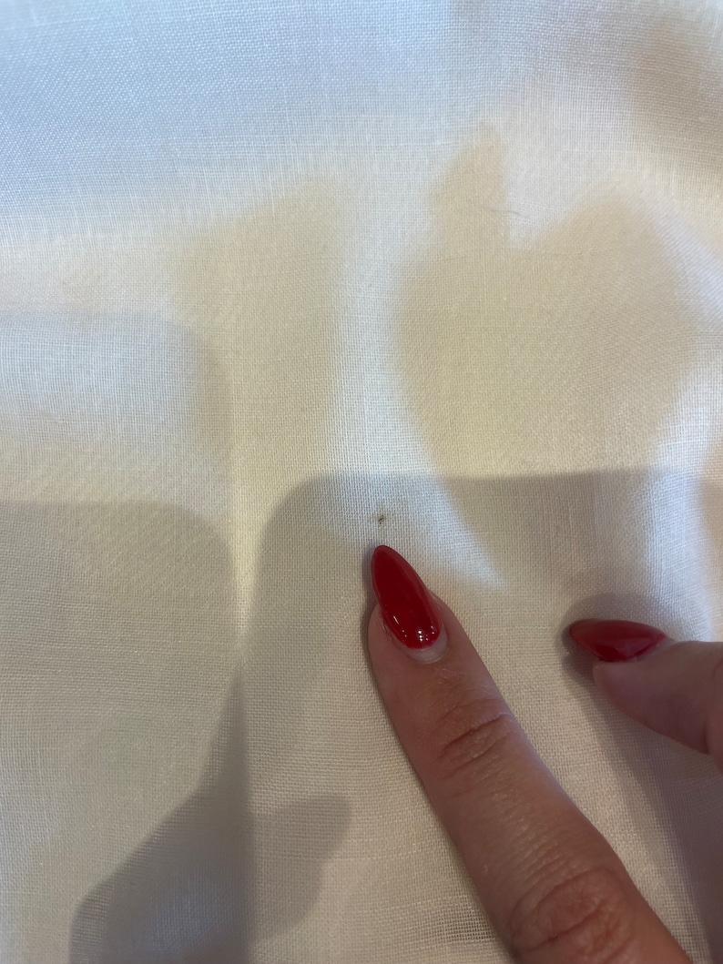May include: A close-up of a white fabric surface with a small brown stain. A finger with red nail polish is pointing at the stain.