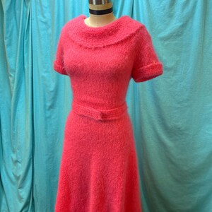 May include: A coral-colored, short-sleeved dress with a wide, ruffled collar and a matching belt. The dress is made of a soft, textured knit fabric and has a flared skirt. The dress is displayed on a mannequin.