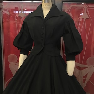 May include: A black dress with a fitted bodice and a full skirt, displayed on a mannequin. The dress features a high collar, button-down front, and three-quarter length sleeves with puffed shoulders. The skirt flares out from the waist.