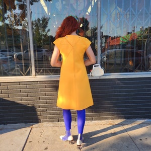 May include: A woman wearing a bright yellow dress with a unique back design, blue tights, and white shoes. She is standing in front of a shop window.