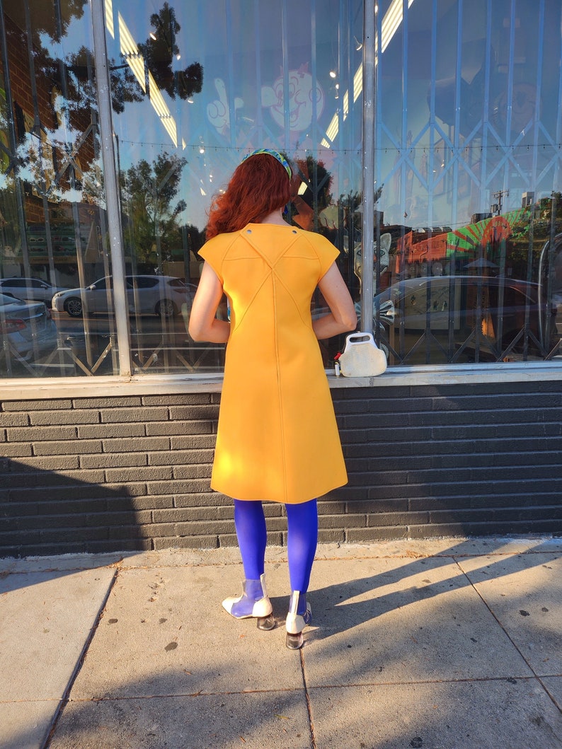 May include: A woman wearing a bright yellow dress with a unique back design, blue tights, and white shoes. She is standing in front of a shop window.