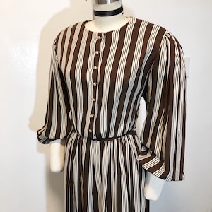May include: A brown and white striped dress with long sleeves and a button-down front. The dress has a fitted waist and a flowing skirt. The sleeves are gathered at the cuffs. The dress is displayed on a mannequin.