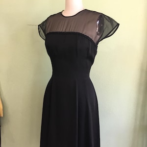 May include: A vintage black dress displayed on a mannequin. The dress features a sheer neckline and cap sleeves, with a fitted bodice and a flared skirt. The dress is a classic style, suitable for formal occasions.