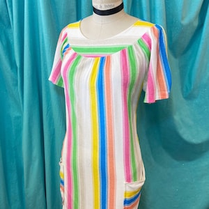 May include: A colorful, striped dress with a scoop neckline and short sleeves. The dress features vertical stripes in various colors, including pink, green, yellow, and blue, against a white background. It has two small pockets and is displayed on a mannequin.