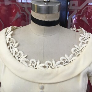 May include: Cream-colored vintage dress on a mannequin. The dress features a wide, rounded collar and a delicate lace trim around the neckline. Small, round buttons run down the front. A black ribbon choker is around the mannequin's neck.