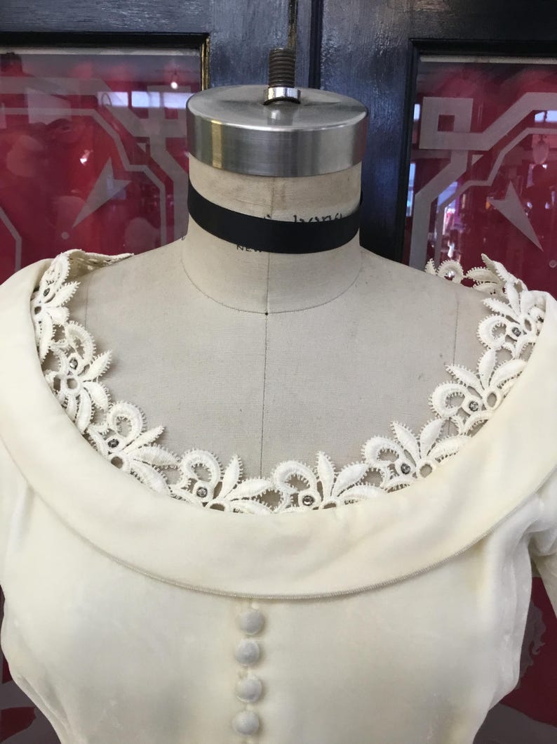 May include: Cream-colored vintage dress on a mannequin. The dress features a wide, rounded collar and a delicate lace trim around the neckline. Small, round buttons run down the front. A black ribbon choker is around the mannequin's neck.