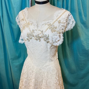 May include: A white lace dress with a scalloped neckline and short sleeves. The dress has a fitted bodice and a full skirt. The lace is embellished with gold sequins and beads.