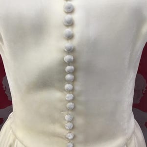 May include: Close-up of a cream-colored dress with a row of small, round, fuzzy buttons running down the back. The dress has a simple, elegant design with a subtle sheen. The buttons are evenly spaced and add a delicate detail.
