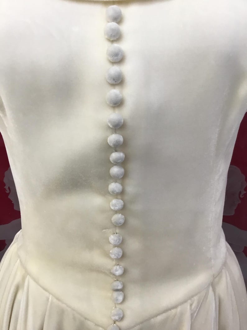 May include: Close-up of a cream-colored dress with a row of small, round, fuzzy buttons running down the back. The dress has a simple, elegant design with a subtle sheen. The buttons are evenly spaced and add a delicate detail.