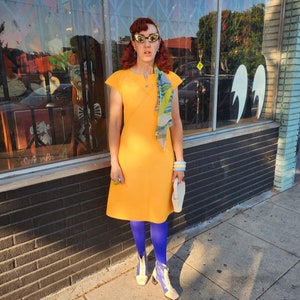 May include: A woman wearing a yellow dress, blue tights, and white boots with a white clutch purse. She is standing in front of a shop front with a glass door and brick wall.