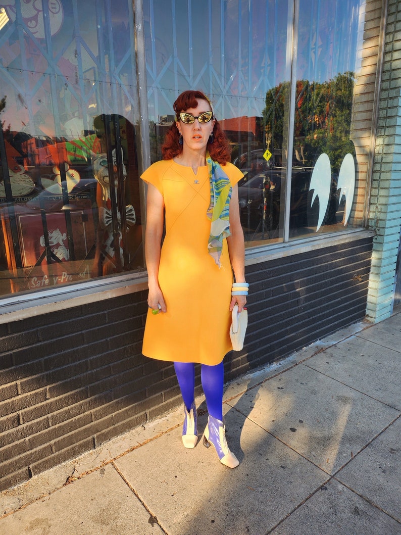 May include: A woman wearing a yellow dress, blue tights, and white boots with a white clutch purse. She is standing in front of a shop front with a glass door and brick wall.