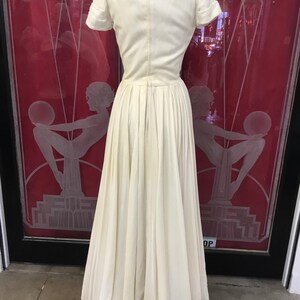 May include: Ivory-colored vintage dress with a fitted bodice and a full, pleated skirt. The dress features short sleeves and a delicate lace collar. The dress is displayed on a mannequin, showcasing its elegant design and classic silhouette. The background includes a red door with etched art deco figures.