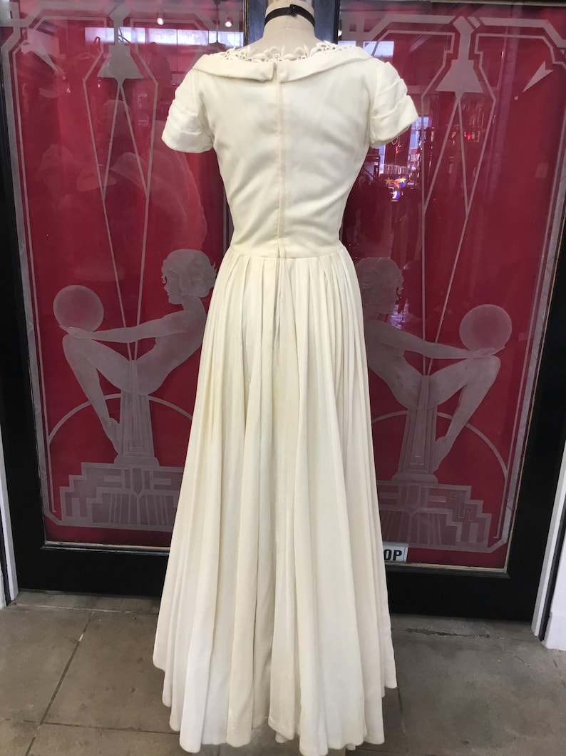 May include: Ivory-colored vintage dress with a fitted bodice and a full, pleated skirt. The dress features short sleeves and a delicate lace collar. The dress is displayed on a mannequin, showcasing its elegant design and classic silhouette. The background includes a red door with etched art deco figures.