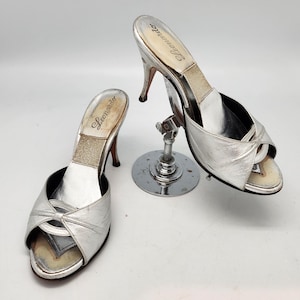 May include: A pair of silver open-toe heeled sandals with a bow detail on the front. The sandals are labeled "Lecenciando" on the insole.